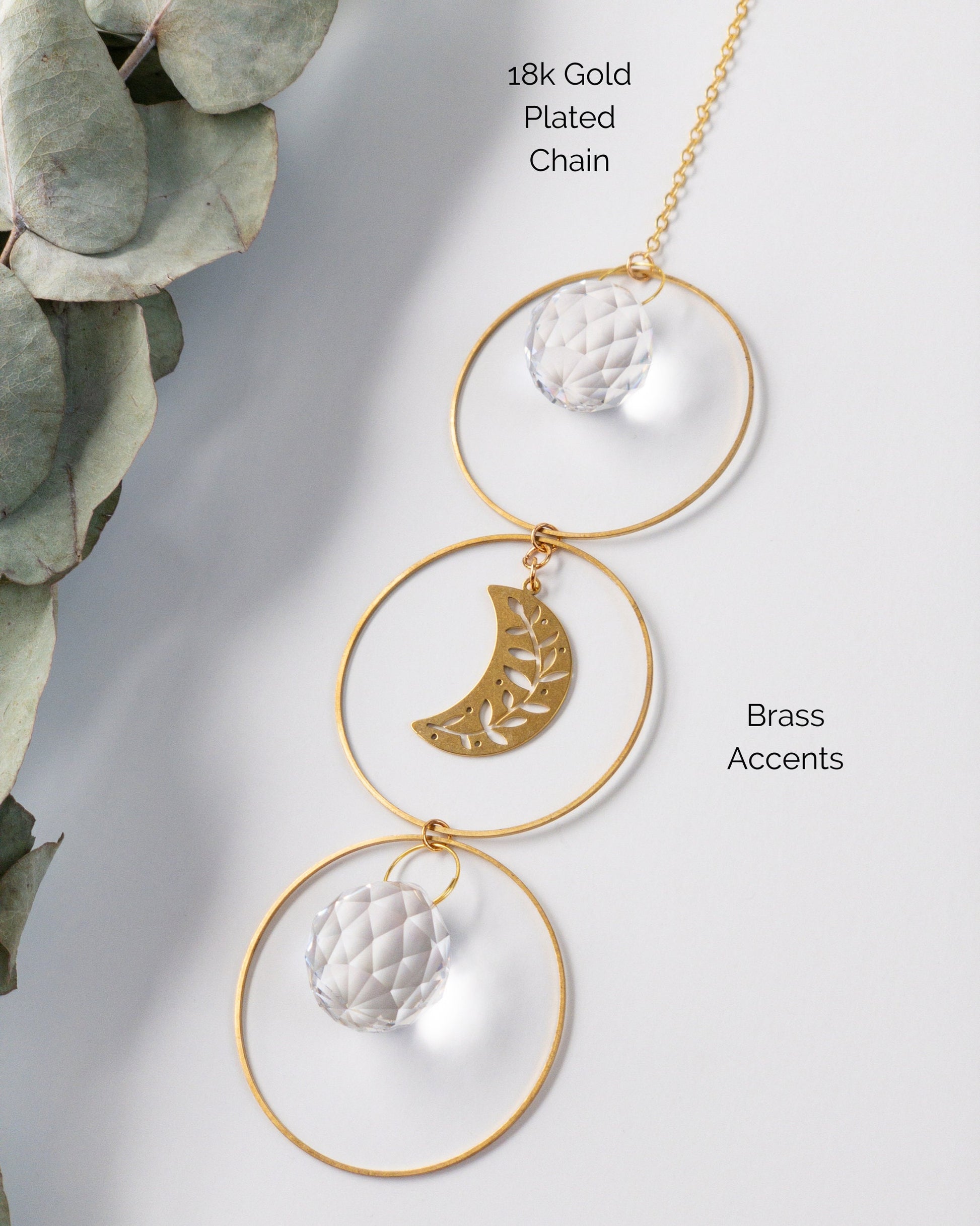 A closeup showing the materials used to make the Brass and crystal suncatcher. Three brass hoops connected vertically. Two Small round crystals. A brass crescent moon with leaf cut outs in the center hoop. 18k gold plated chain.