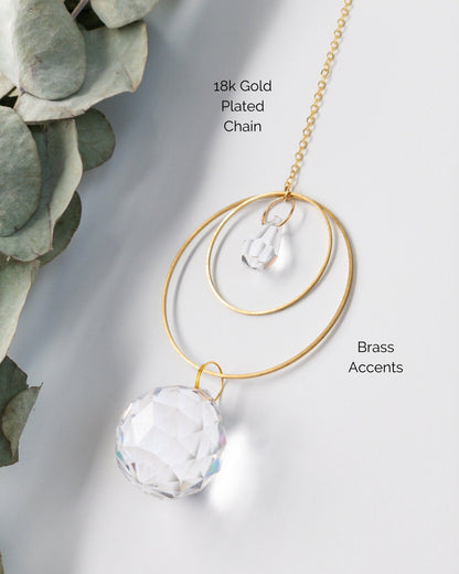 A closeup showing the materials used to make the Brass and crystal suncatcher. Two brass hoops, one small and one large. Small teardrop crystal inside the small hoop. A larger round crystal ball hangs below the large hoop. 18k gold plated chain