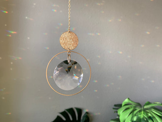 Brass and crystal suncatcher that disperses rainbow flecks around the walls and floor. A brass hoop is suspended from a textured brass circle. A large disc shaped crystal is suspended in the middle of the hoop.