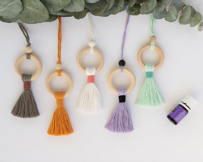 Decor piece to hang from the rearview mirror of your car. Features a wood ring with colorful brushed macrame cord tassel. Some have a wood bead at the top of the ring, others have a black or white lava bead. A bottle of essential oils in the corner.