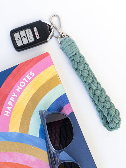 Wristlet Style Teal Macrame keychain in a braided pattern with key fob attached. Styled with a notebook and sunglasses.