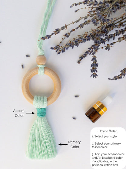 Decor piece to hang from the rearview mirror of your car. Features a wood ring with colorful brushed macrame cord tassel. A wood bead at the top of the ring. A small dropper bottle of essential oils in the corner.