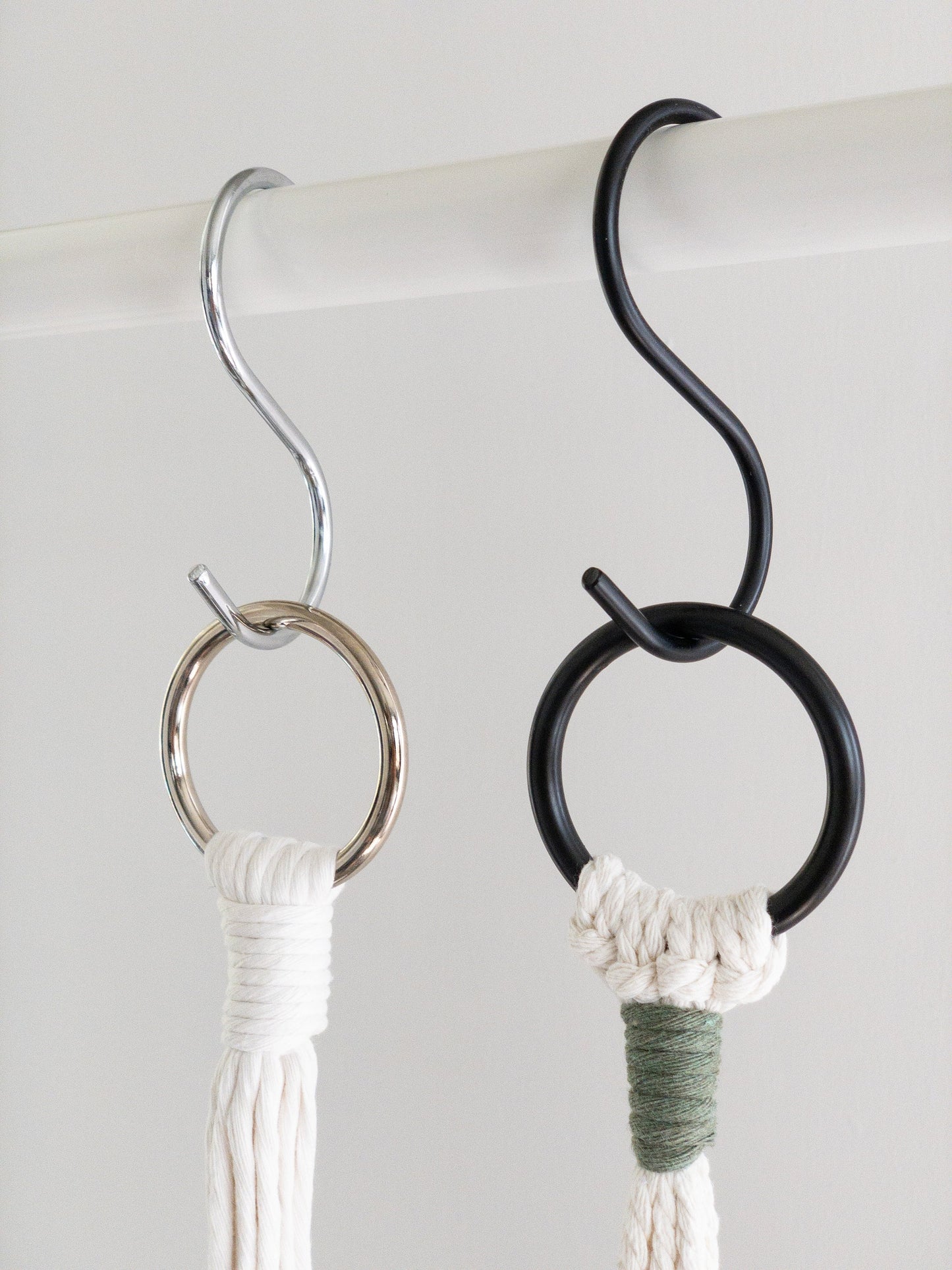 A white rod with two metal S shaped hooks hanging. The S hook on the left is silver and the S hook on the right is matte black. A plant hanger is attached and hanging from each S hook.