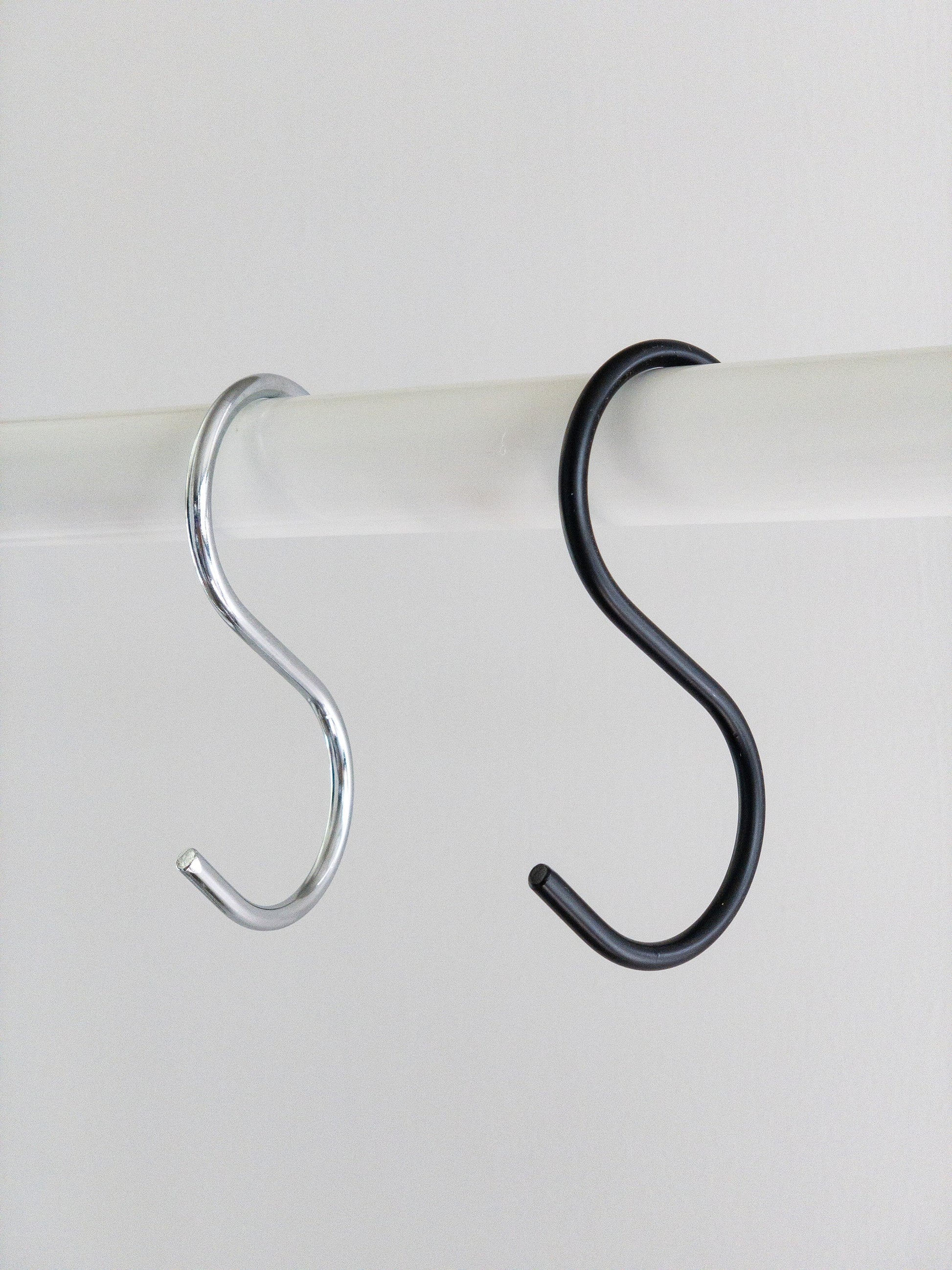 A white rod with two metal S shaped hooks hanging. The S hook on the left is silver and the S hook on the right is matte black.