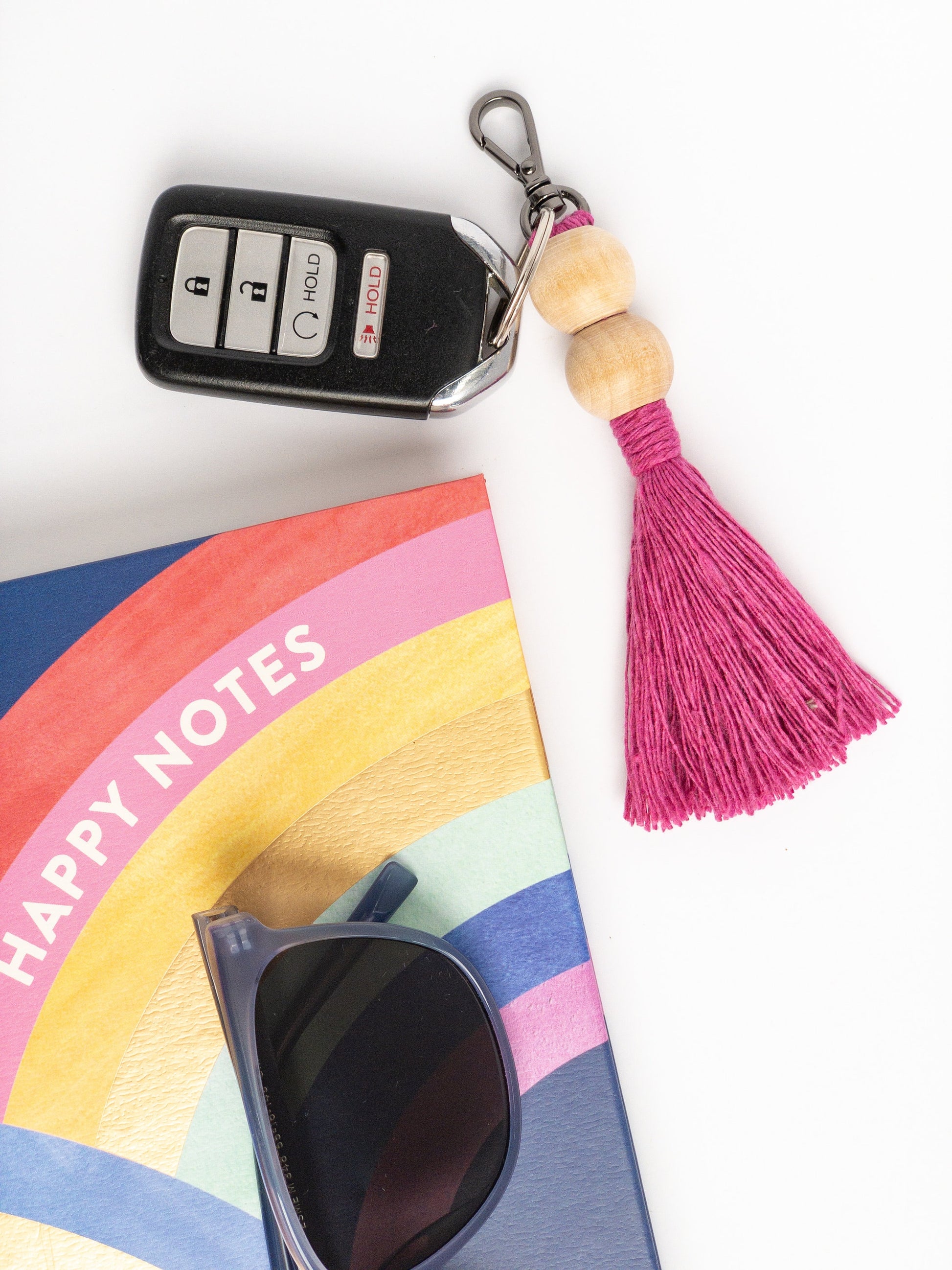 Keychain attached to a keyfob featuring two round beads at the top with a colored tassel (hot pink) below the beads. Styled with sunglasses and notebook.