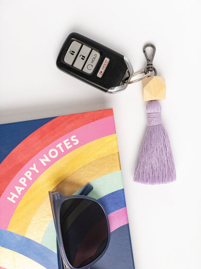 Keychain attached to a keyfob featuring 3 dimensional wood hexagon bead at the top with a colored tassel (lavender) below the beads. Styled with sunglasses and notebook.