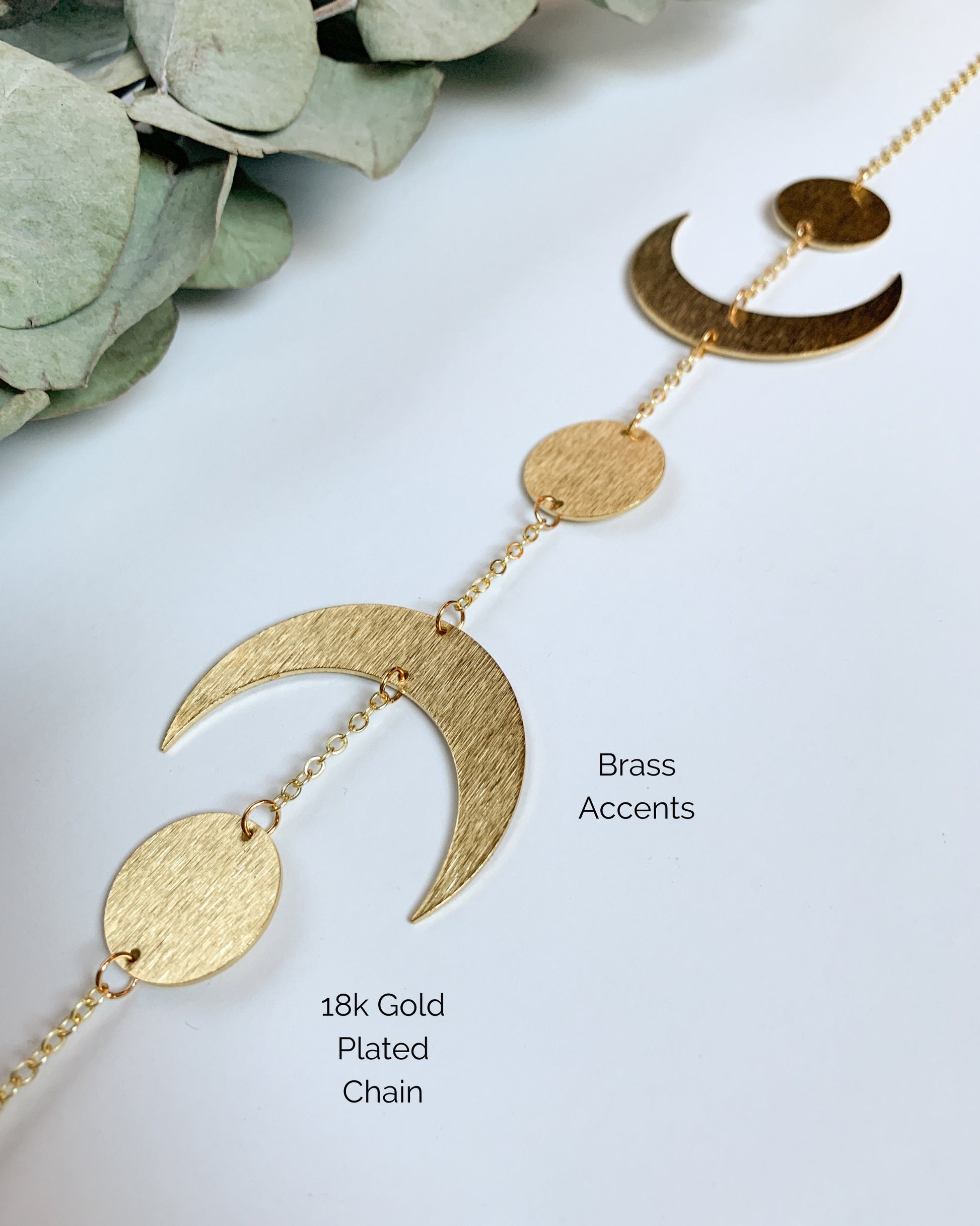 A closeup showing the materials used to make the Brass and crystal suncatcher. Two textured brass crescent moons shapes and three textured brass round shappes. A large round crystal ball hangs from the bottom. 18k gold plated chain.