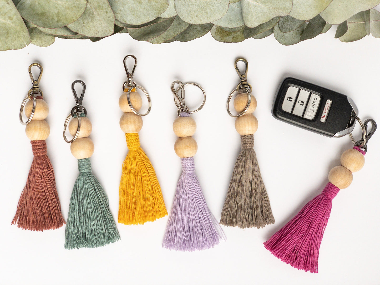 Macrame keychain shown in 6 colors. Brass, silver and smoke colored hardware. Two round beads at the top with a colored tassel below the beads. Keyfob shown on one keychain. Rosewood, Teal, Yellow, Lavender, Taupe and Hot Pink examples.